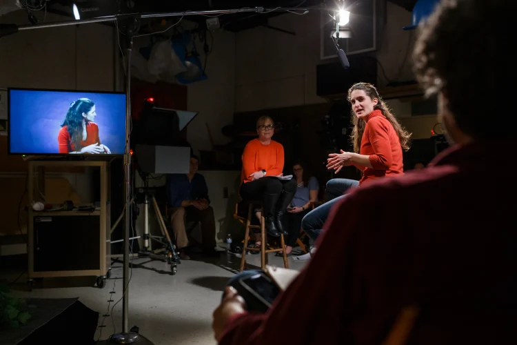 A reporter and interview subject are seen sitting on stools with a microphone, studio camera equipment, and a live monitor showing a video feed of the interview subject.