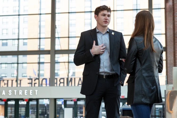 A man gestures at his chest with his hand as he stands facing a woman in conversation. A wall of windows and downtown buildings are seen in the background.