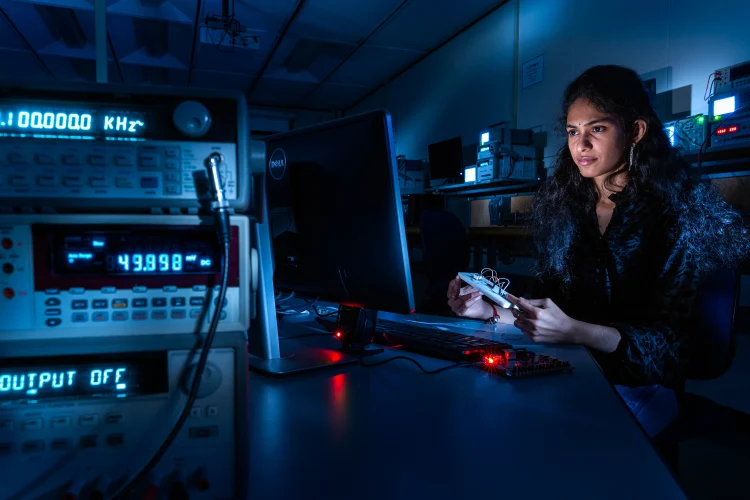 A student in holds a piece of wired technology about the size of her hand while looking at a computer monitor in dark lab setting. Nearby electronic appliances show readings of numbers on their screens.