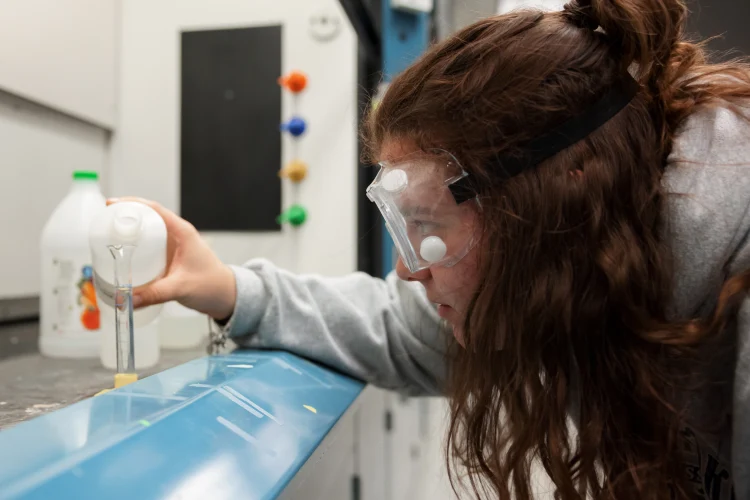 A female student wearing protective goggles leans over into a ventilated lab area to pour a solution into a test tube in a stand.