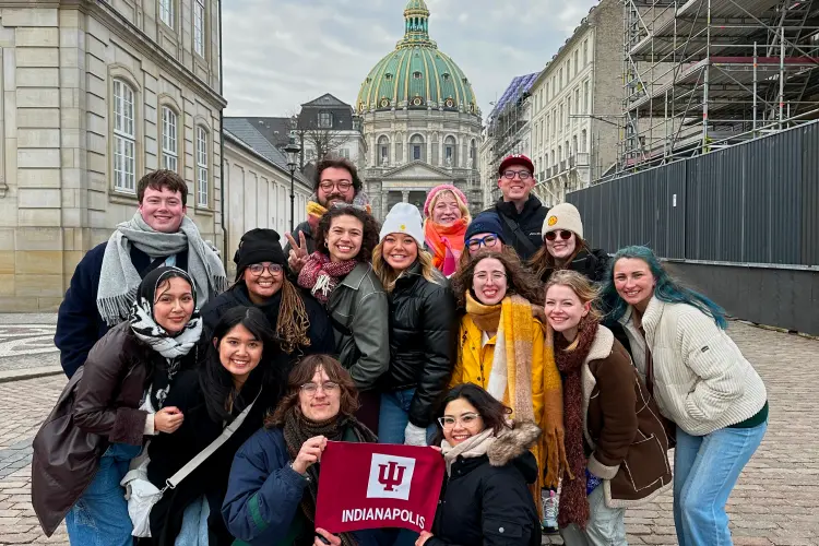 A group of about 16 IU study abroad students gathers on a brick street with the front two kneeling and holding a small IU Indianapolis flag. A domed stone building is seen behind them at the end of the street.
