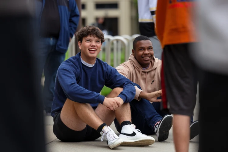 Two male students sit on the ground outdoors during a Bridge Week group activity.