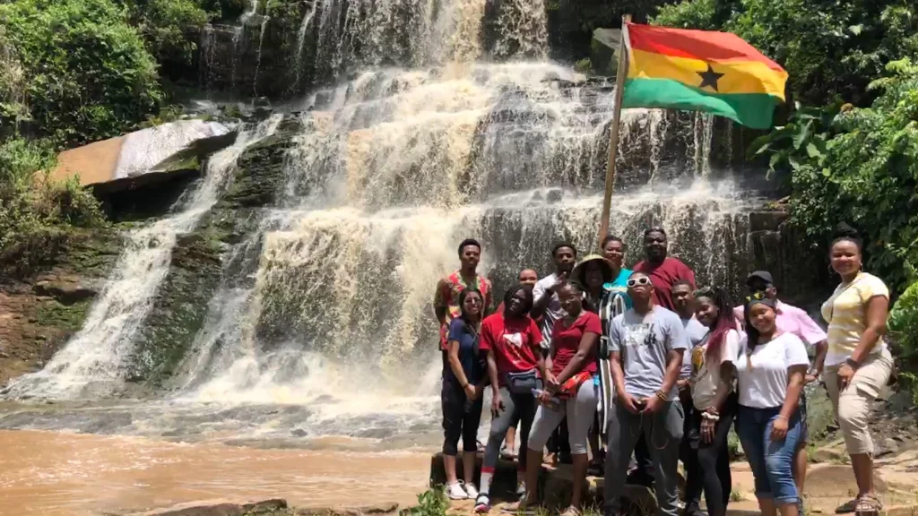 IU Indianapolis students gather in a group on a rocky shore in front of a multi-layered waterfall and country flag blowing in the wind in Ghana.