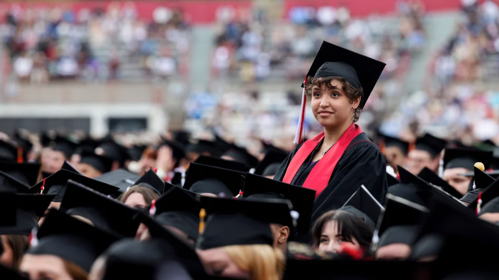 A graduate in cap and gown stands up, surrounded by other graduates seated at Carroll Stadium for Commencement ceremony. Attendees are seen out of focus in stands in the background.