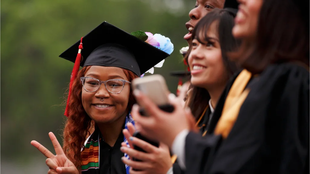 A student leans out from a row of graduates wearing caps and gowns, smiling and holding up two fingers.