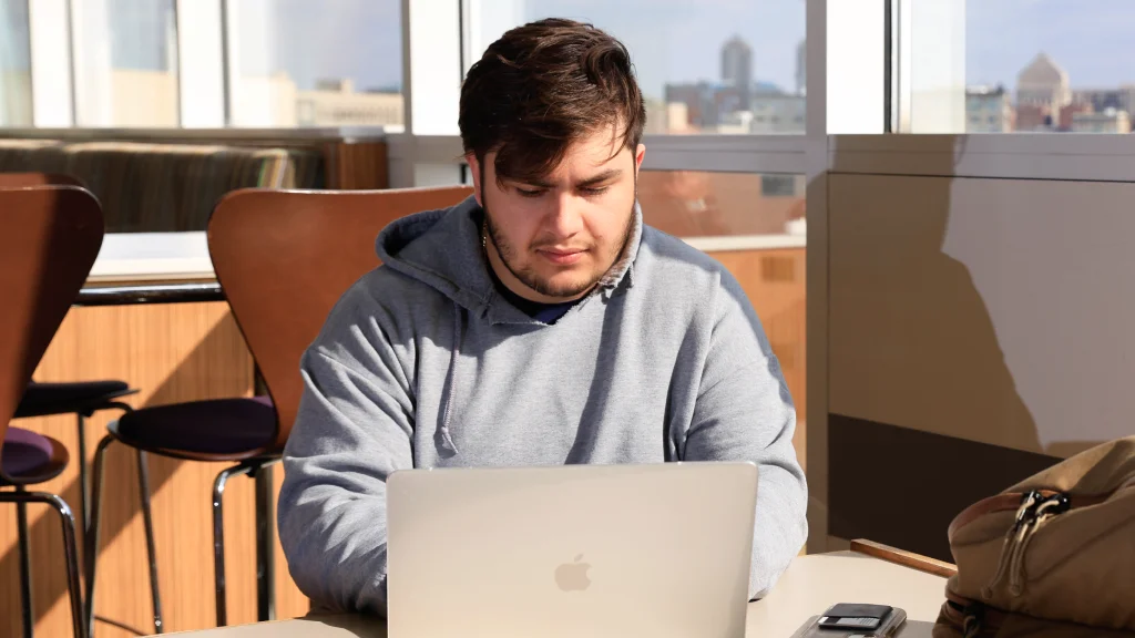 A student sits at a table with an open laptop, backpack nearby, at a community space in University Library with windows overlooking downtown Indianapolis.