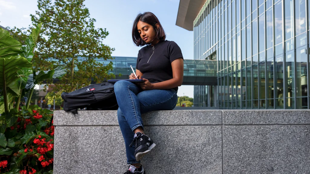 A student sits on a high marble wall; flowering plants are seen to the left and the glass of the Campus Center facade and a pedestrian skywalk are seen behind her and to the right.