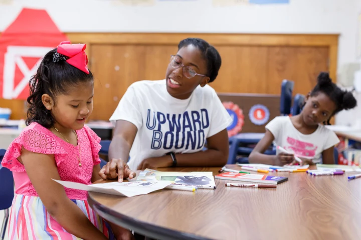 A teacher sits between you young girls in a classroom pointing to a piece of paper in front of one. The other draws with colored markers.