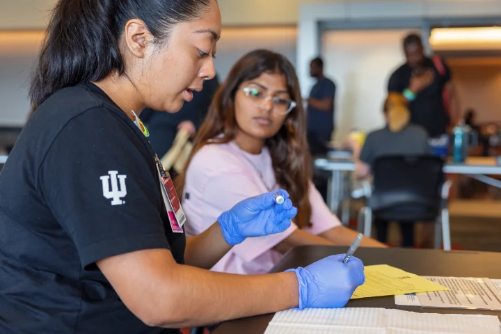 A woman sits at a table in a large room wearing a tee shirt with a trident on the sleeve. She wears medical gloves and holds a vaccine in one hand and a pen in the other, writing on paperwork. A girl sits nearby in a chair watching as the vaccine is prepared for injection.