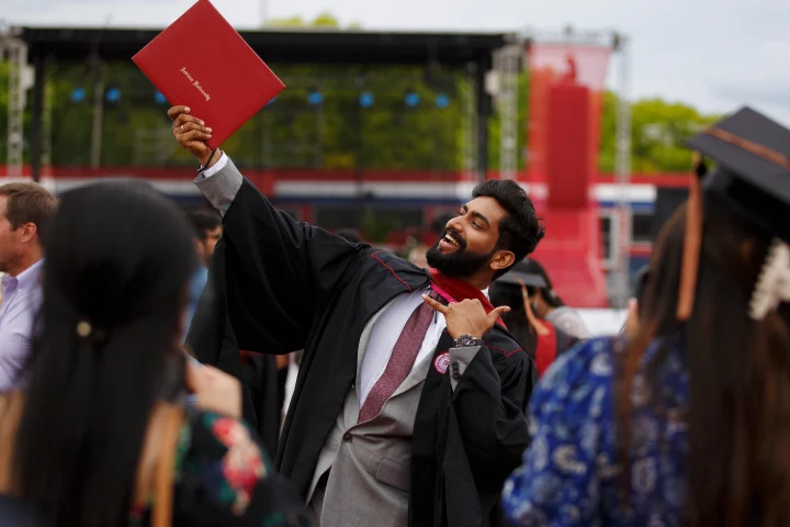 A graduate student in Commencement gown holds up a red IU diploma cover in the air in celebration; the ceremony stage is seen in the background.