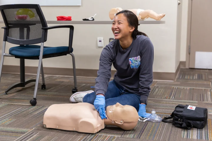 A student sits on a carpeted floor feeling for a pulse on a dummy mannequin as she practices CPR and emergency response techniques.