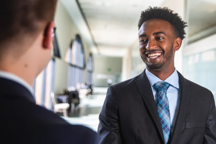 Two students in suits speak with each other in a large, airy, sun-filled hallway.