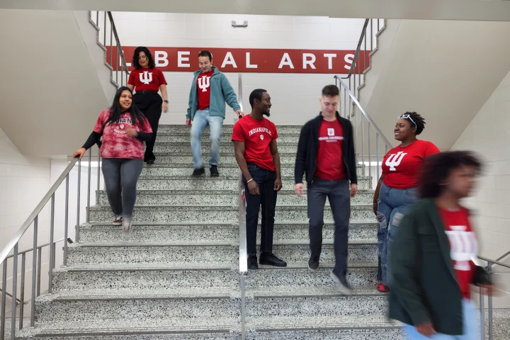 Seven students, many wearing crimson IU shirts, stand and walk on an interior marble double staircase. A large wall sign on the top landing of the stairs says Liberal Arts.