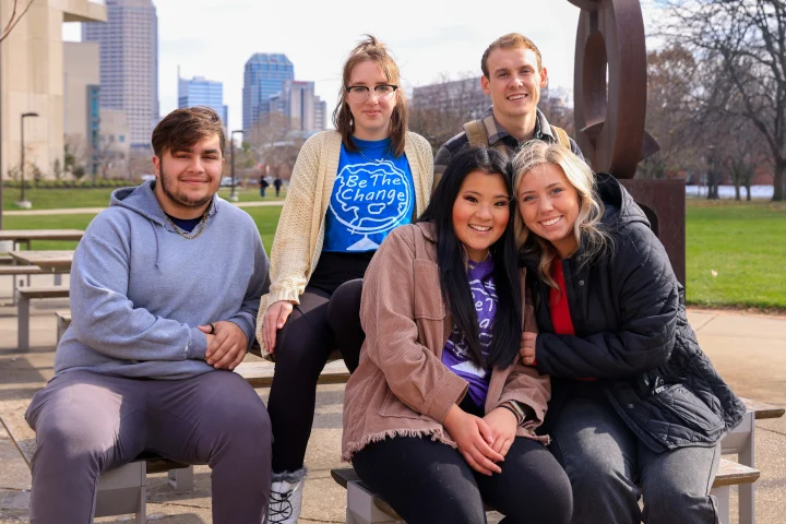 A group of students, two wearing School of Philanthropy shirts that say Be the Change in a globe icon, sit on an outdoor bench on IUI campus with the Indianapolis skyline visible in the background.