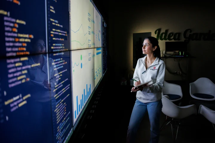 A woman holds a notebook in one hand, pen in the other, as she looks up at a wall screen display of graphics and charts in a darkened room.