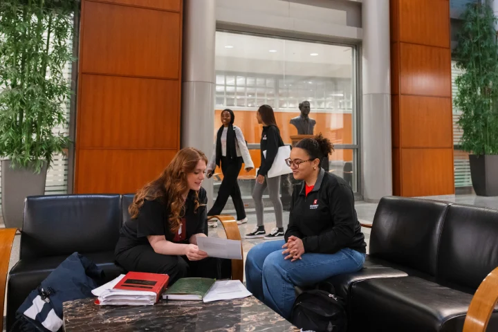 Two law students sit on leather couches in a high-ceilinged common area with law textbooks on a table in front of them.