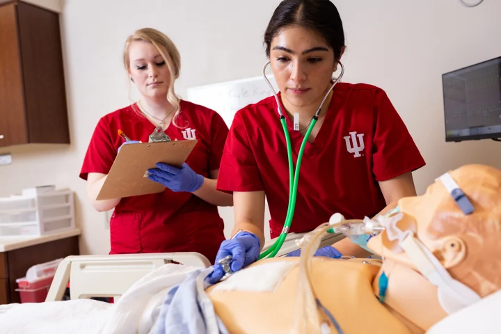 Two nursing students stand behind a dummy patient in a simulation lab. One student holds a stethoscope to the dummy’s chest, the other stand behind writing on a clipboard.