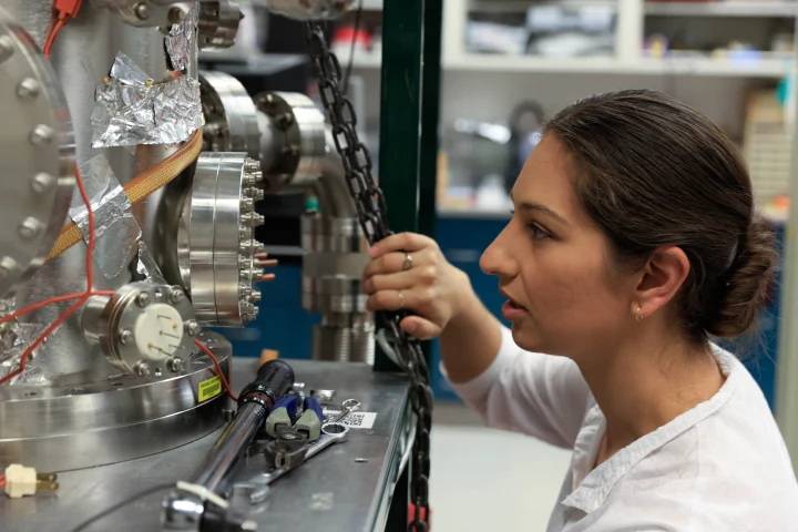 A woman kneels down to look closer at a metal piece of machinery. Tools sit on a table in front of the piece of machinery.