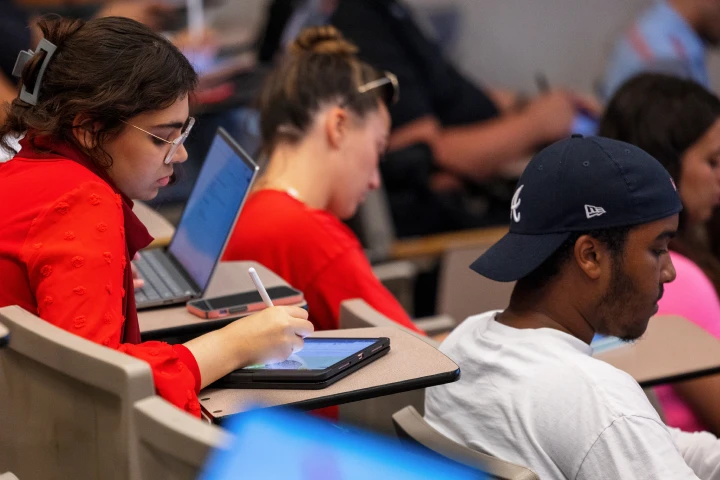 A girl in a red sweater sits in a lecture hall seat writing on a tablet. Several students fill the seats around her for a class.
