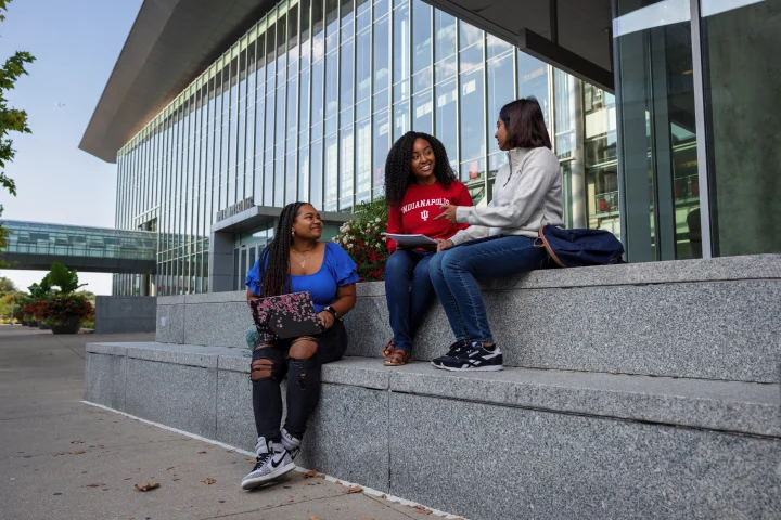 Three students sit on wide stone stairs in front of the Campus Center’s glass facade.