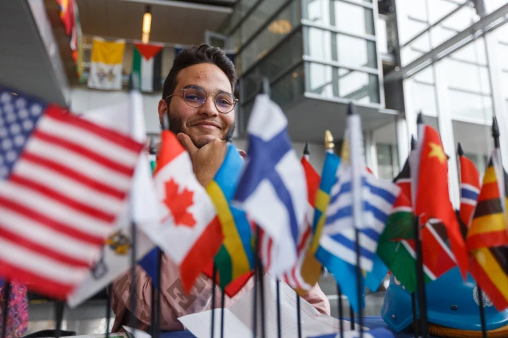 A student smiles. chin on hand, as he sits behind a tabletop display of international flags during an event in the Campus Center atrium.