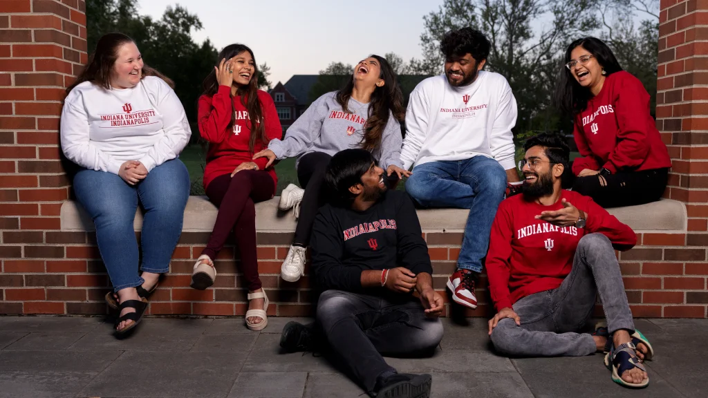A group of seven students wearing IU Indianapolis shirts sit on or in front of a low brick wall on campus chatting and laughing with each other.