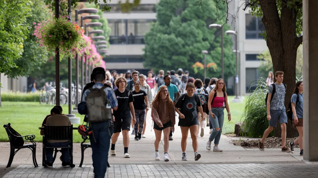 Dozens of students walk along a campus sidewalk lined with light poles adorned with hanging flower baskets. One student sits at on outdoor table and a bike rack is seen in the distance.