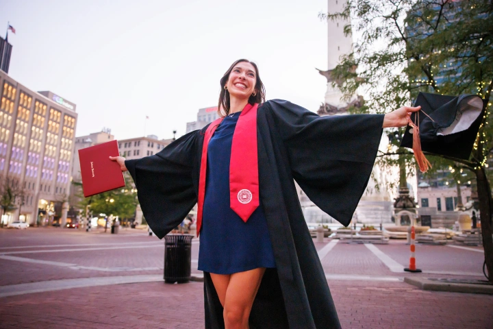 A female graduate wearing a dress covered by a commencement gown holds a diploma cover in one outstretched hand and mortarboard in the other outstretched hand. She stands on the bricks on Monument Cirle, with buidlings and the monument visible in the background.