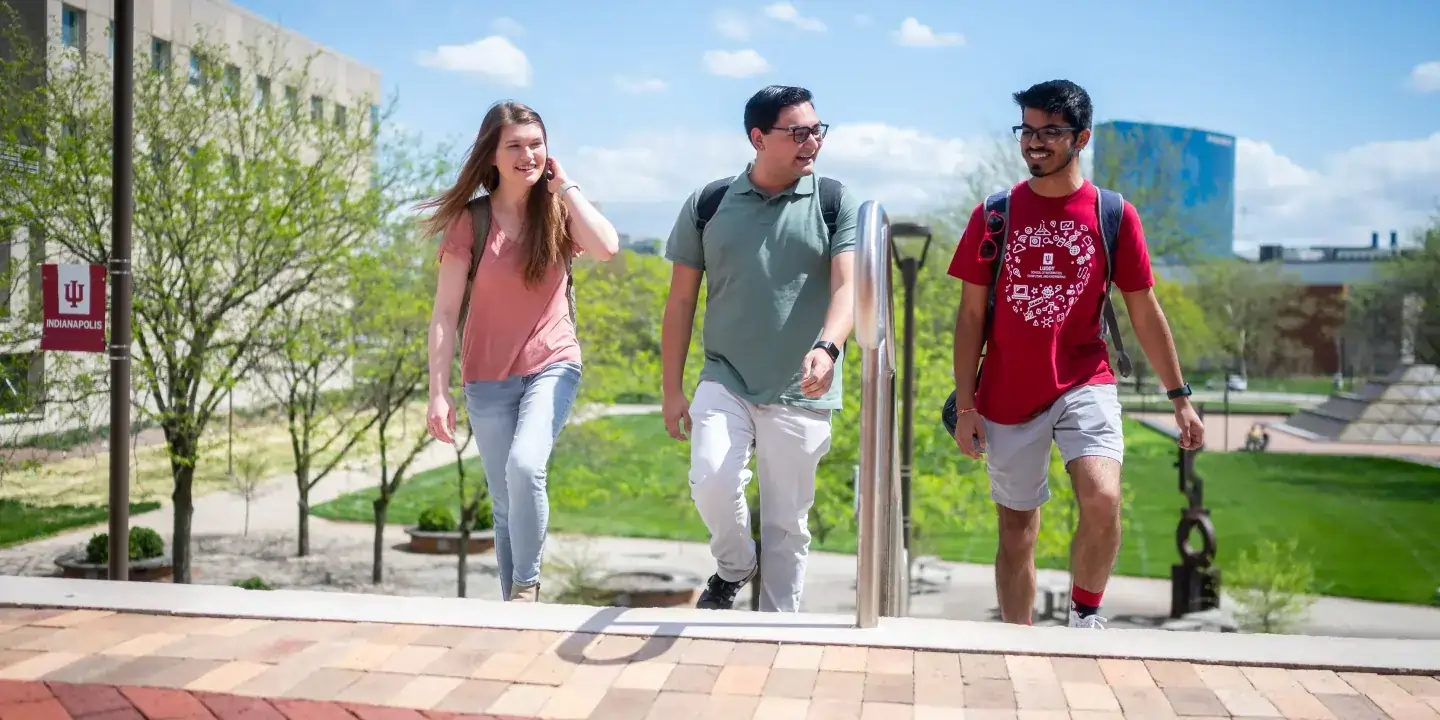Three students walk to the top of a bricked stairway, each sporting a backpack. Green blooming trees dot the lawn behind them, with the University Library on the left and the skyline of Indianapolis rising to the right.