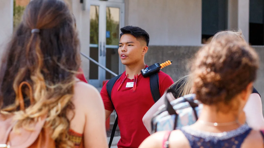 A student tour guide in an OTEAM shirt with walkie talked on his backpack strap faces tour attendees in front of a building on campus.