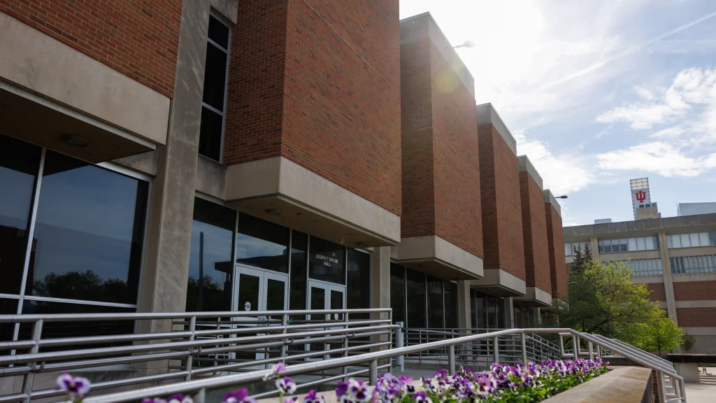 Brick building with vertical windows inset from the facade, flowers blooming in an elevated landscaped area in front of entrance ramp and stairs.