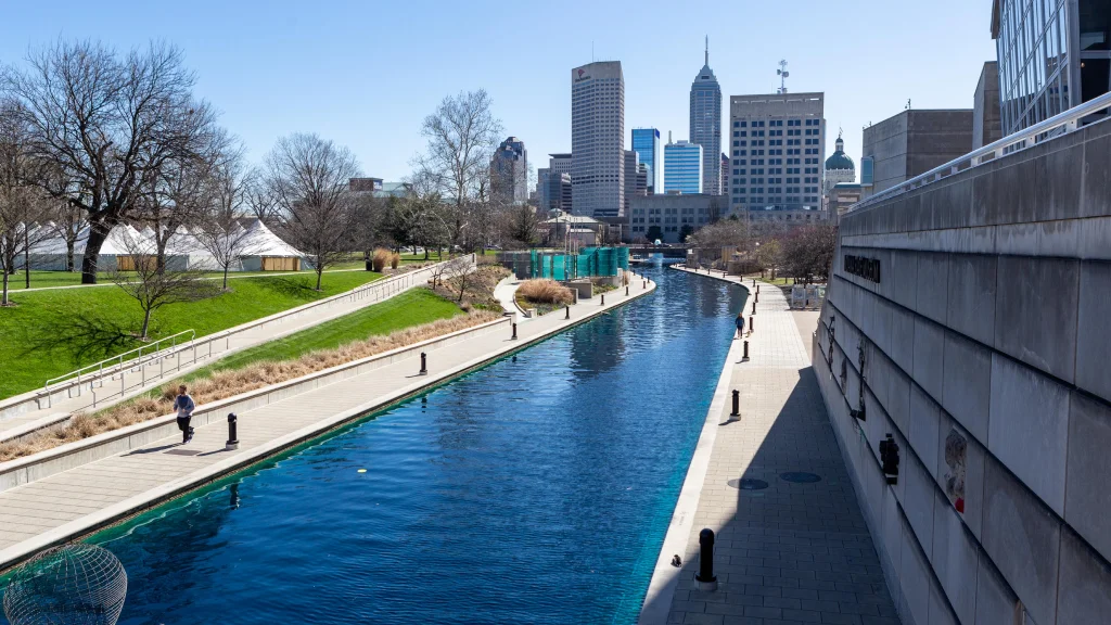 Scenic canal with walkways on both sides, bordered by green lawns and trees, with the Indianapolis city skyline in the background.