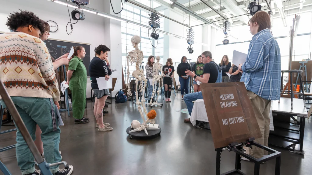 Art students gathered around skeleton models in a well-lit studio with large windows and high ceilings.