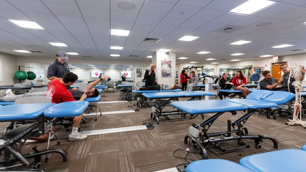 Students practicing physical therapy techniques on treatment tables in a large clinical classroom.