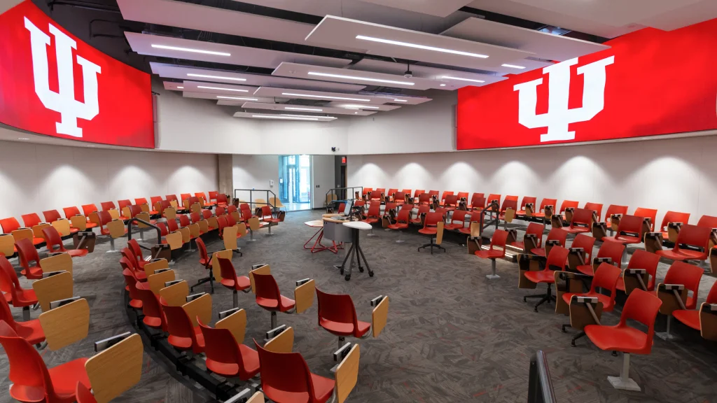 Modern classroom with rows of bright red chairs with attached desks arranged in a circle around a center lectern. IU logos are displayed on large screens on the walls above the chairs.