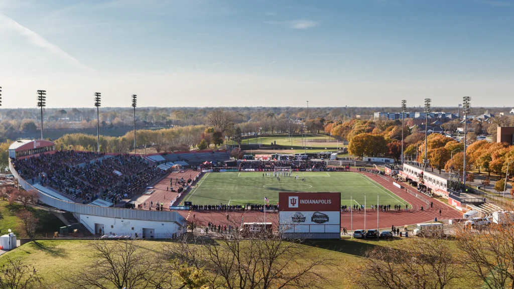 Large outdoor stadium filled with spectators, surrounded by trees with autumn foliage.