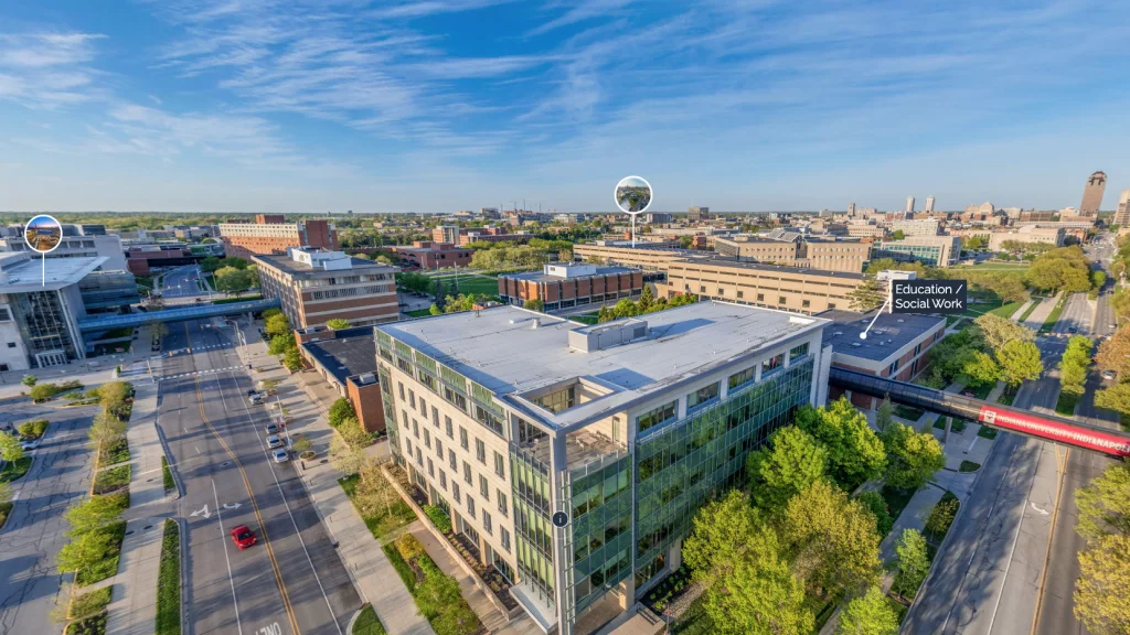 Aerial view of modern university buildings with large glass windows, situated on tree-lined streets with adjacent academic buildings and green spaces and the city skyline visible in the distance.