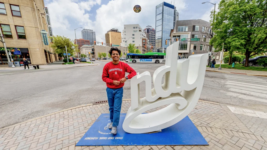 Smiling person in a red Indiana University sweatshirt standing next to a large "Indy" sculpture on a city street with buildings, trees, and a bus in the background.