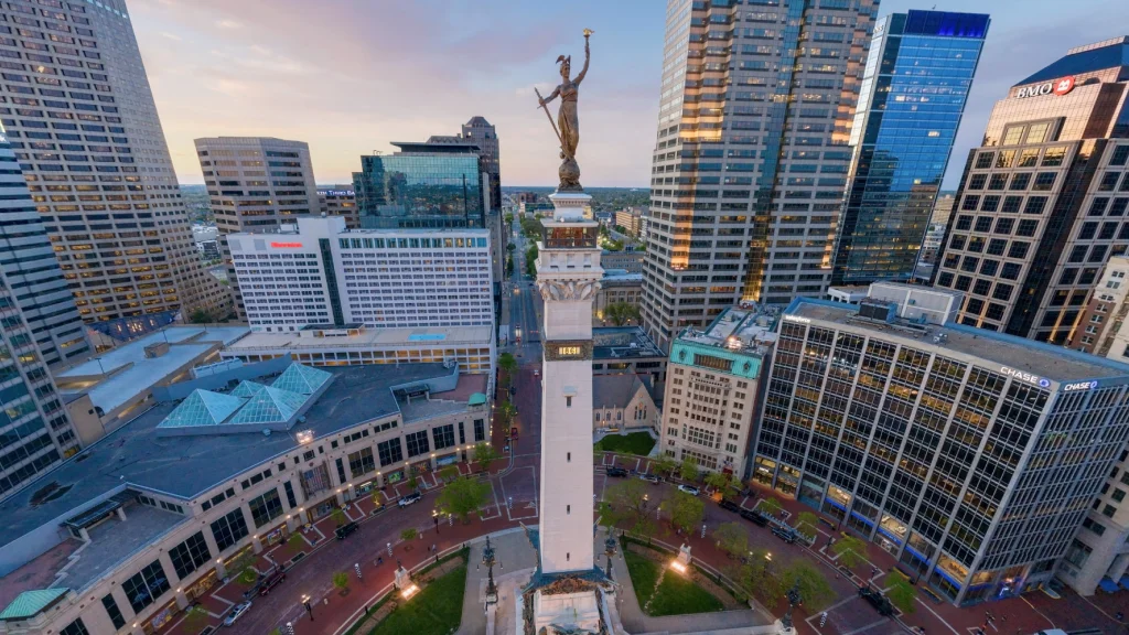 Aerial view of downtown Indianapolis at dusk, showcasing the iconic Soldiers and Sailors Monument with its detailed sculpture at the top, surrounded by illuminated skyscrapers.