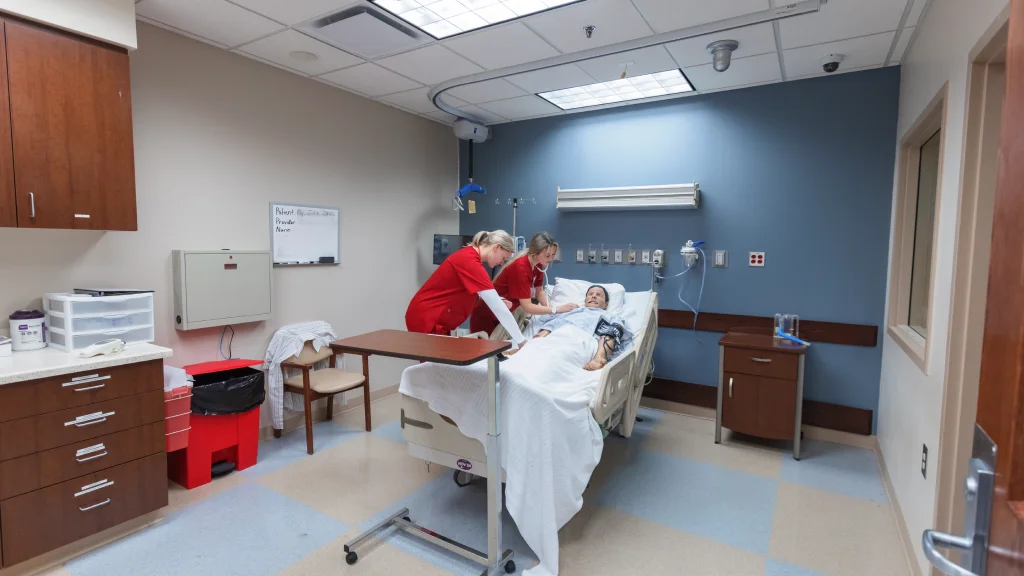 Nursing students in red scrubs practicing on a patient simulator in a clinical training room.