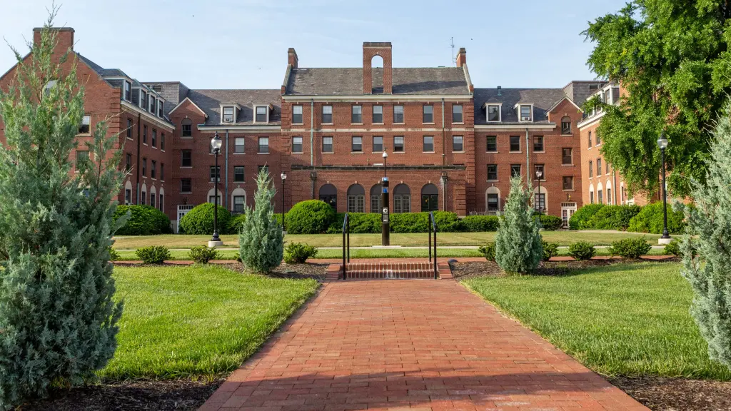 Four story red-brick university building with trees and landscaping in the foreground, with a paved brick sidewalk leading toward the building.