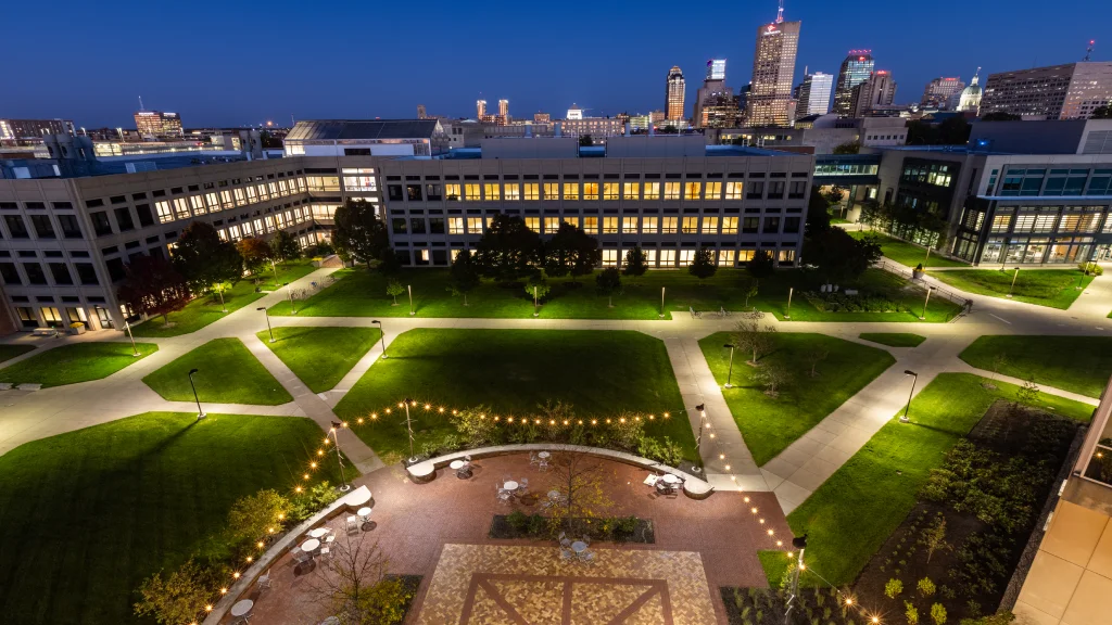 Illuminated university courtyard with walkways and buildings, set against a backdrop of a city skyline.