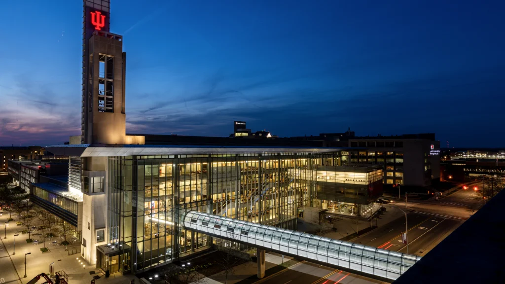 Modern building with a skywalk and illuminated IU logo on a tower.