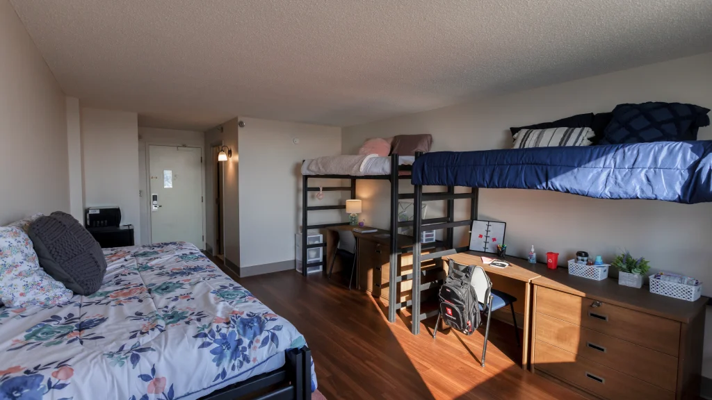 Neatly arranged dorm room with a lofted bed, a desk underneath, and another bed with floral bedding, featuring a wood floor and bright window lighting.