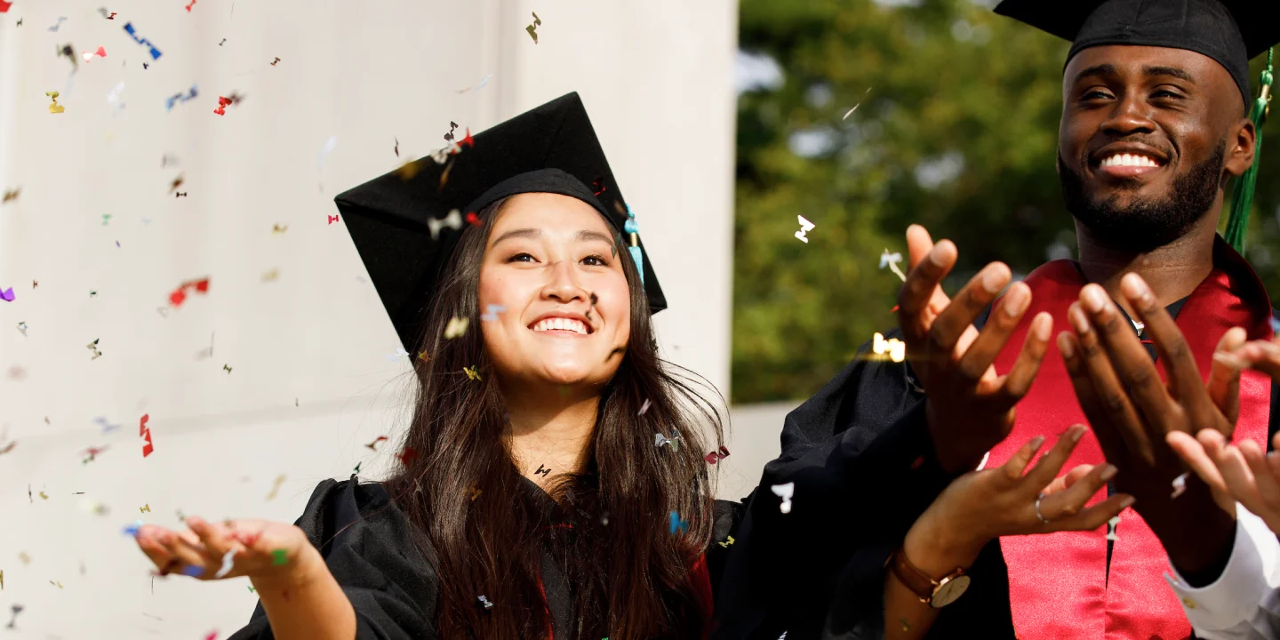 A male and female graduate hold out their hands and look up as colorful confetti falls down around them.