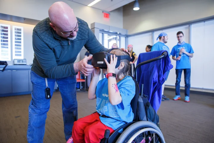 A man adjusts a VR headset around the head of a young child sitting in a wheelchair.