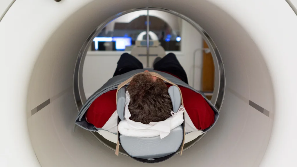 A man is seen laying inside an MRI machine; his head visible and his body loosely wrapped in a blanket-like item.