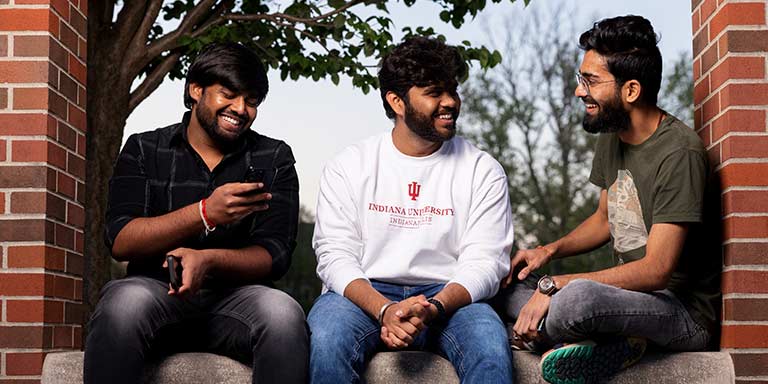 Three male students sit on a brick wall while smiling and laughing at each other, the center student wears an IU Indianapolis sweatshirt.