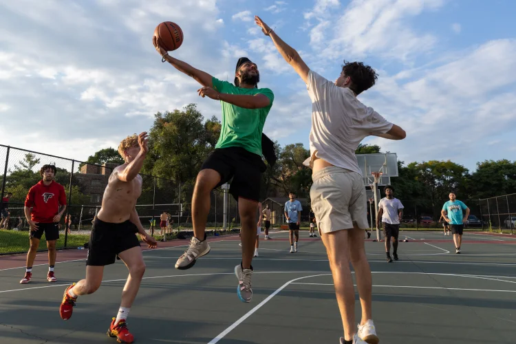 A student jumps up, basketball in hand, while others defend in a game of basketball on an outdoor court.