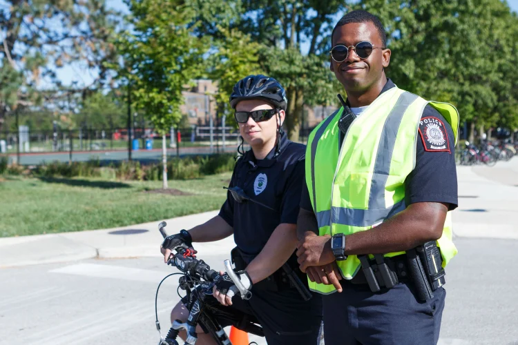 A police officer standing in a neon green and a second police officer seated on a bicycle stand stand side by side in a parking lot.